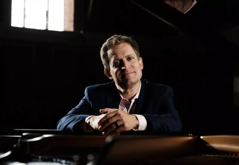 Andy Armstrong with hands clasped over the edge of his grand piano looks softly with a smile into the camera. 