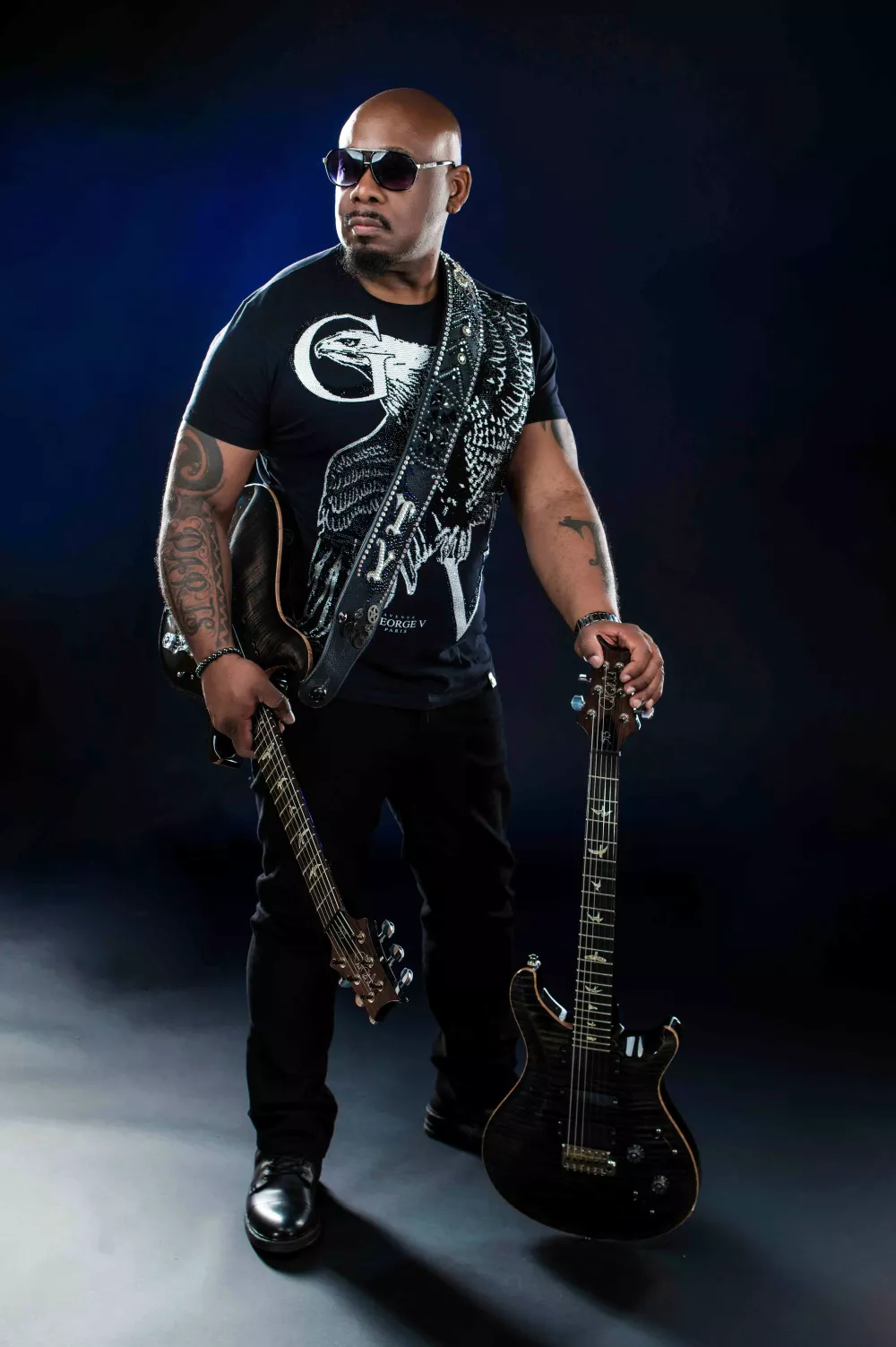 Promotional image of Terence Young wearing one guitar around his chest and one propped on the ground.