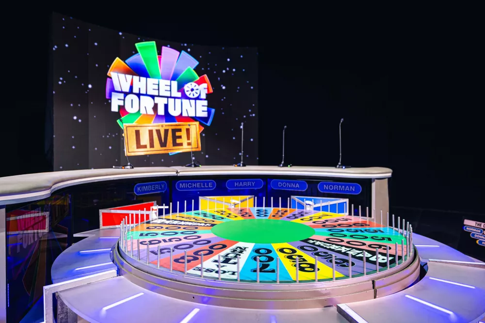Wheel of Fortune LIVE! stage featuring the colorful prize wheel, contestant podiums, and a large screen displaying the show's logo.