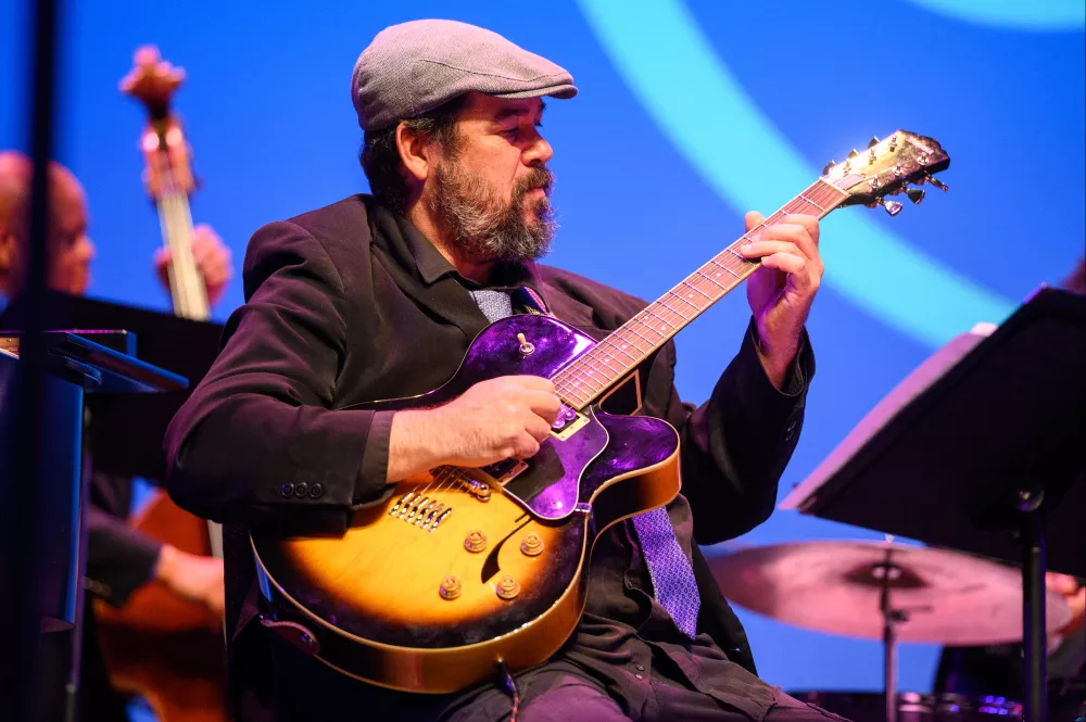 Mna holding a guitar, looking at a a music stand with an upright bass player in the background.