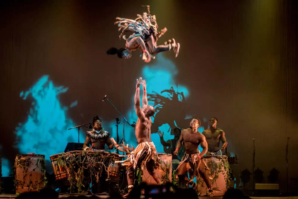 One performer with arms outstretched up as if to catch another performer flipping in the air. Other performers are in the background watching and playing drums.