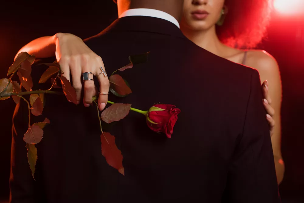 Woman with an arm around a man wearing a tuxedo, with a red rose in her hand.