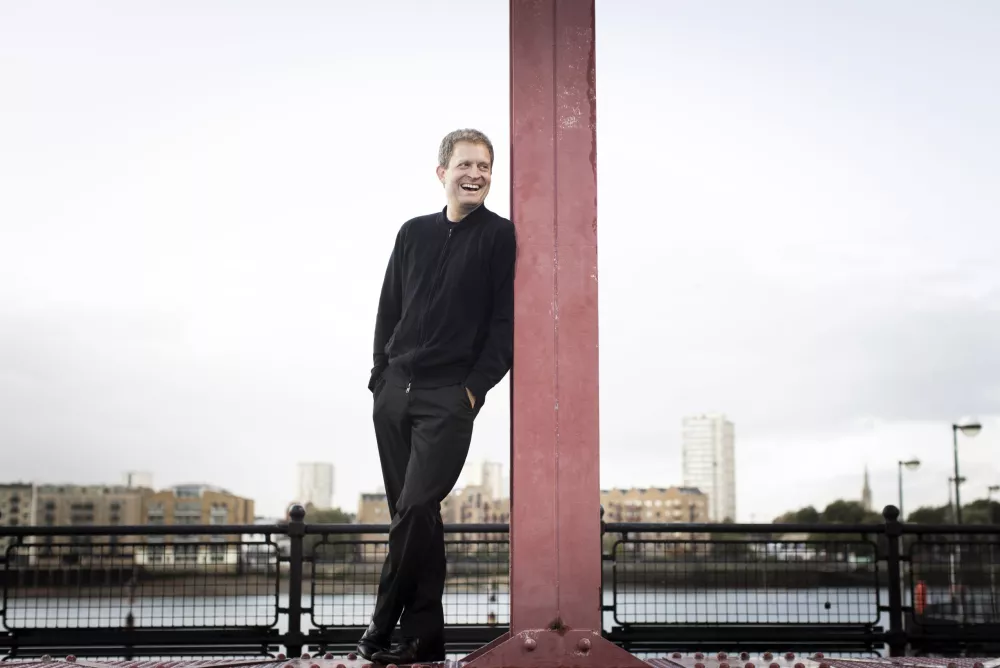 Man wearing black, leaning against pole outdoors and laughing.