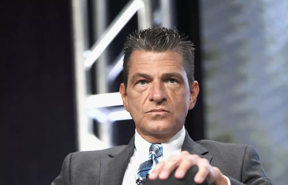 Man in a grey suit and blue tie sitting in a chair with one hand on a knee.