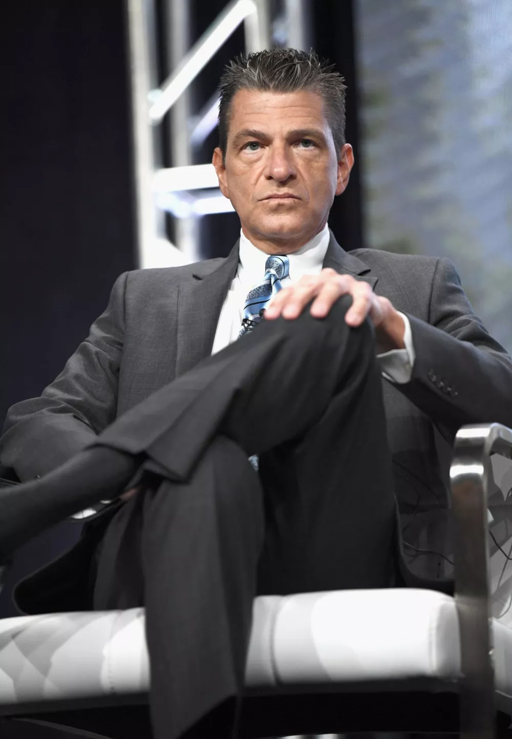 Man in a grey suit and blue tie sitting in a chair with one hand on a knee.