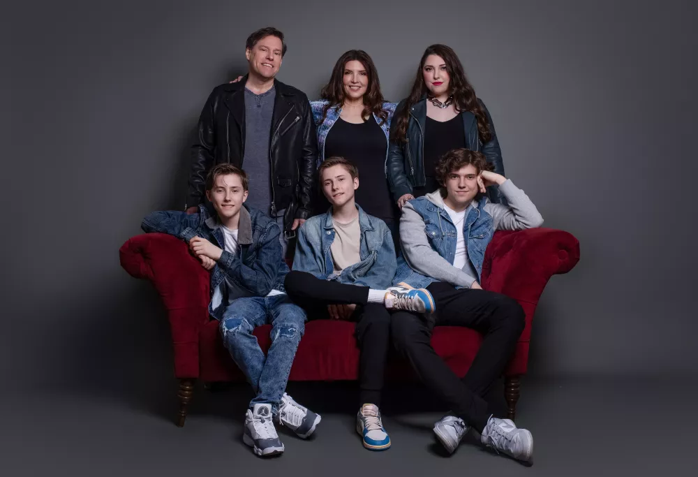 A family of 6 with a father, mother, and daughter standing behind a red couch with 3 sons sitting on it.